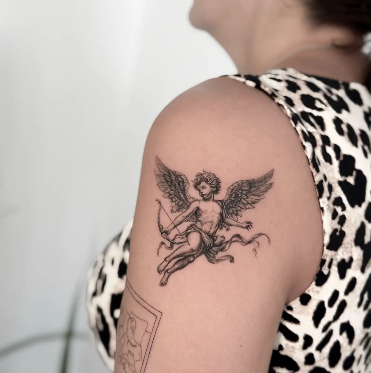 Querubín alado con arco y cinta tatuado en microrrealismo negro y gris sobre el hombro