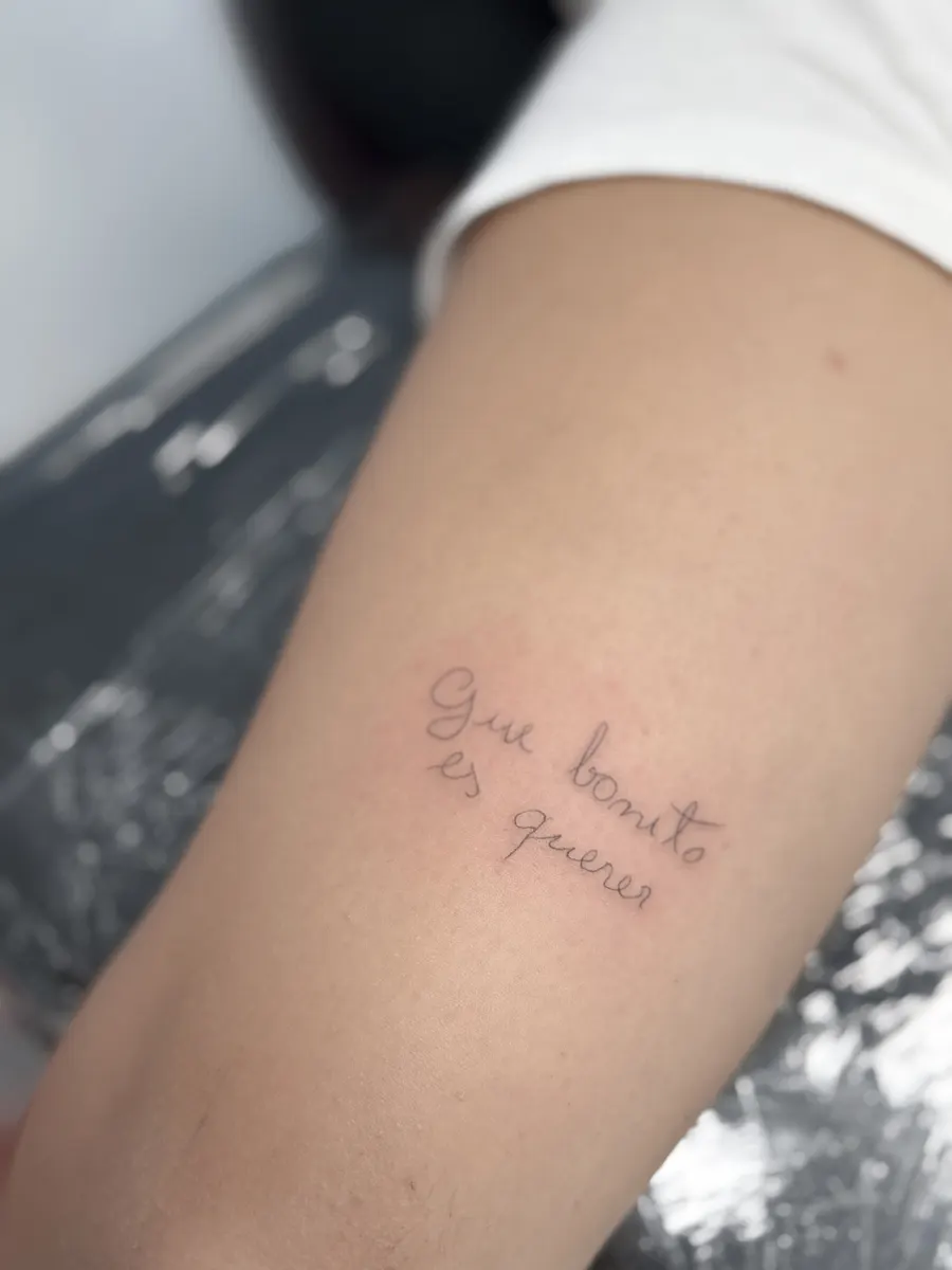 Frase "Que bonito es querer" tatuada en caligrafía manuscrita finísima en el brazo